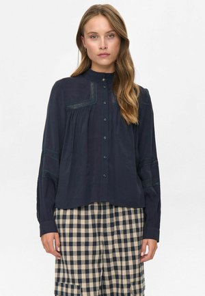 Kvinna iklädd en marinblå blus med knappar och spetsdetaljer samt byxor i beige och marinblå rutig mönster framför en enfärgad ljus bakgrund.