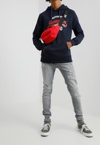 Sudadera navy con gráfico en blanco y rojo, jeans grises desgastados, zapatillas verdes y un bolso cruzado rojo. Atuendo urbano casual.