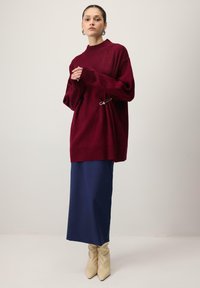 Pull oversize bordeaux avec un col montant et un détail d'épingle de sûreté, associé à une jupe bleu marine et des bottes beige décontractées. Texture lisse sur l'ensemble.