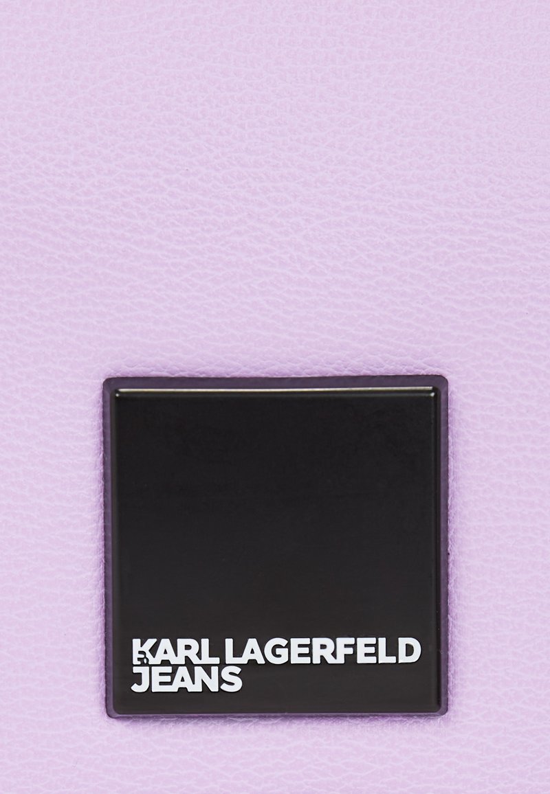 Cuir texturé lavande avec une étiquette carrée noire mate affichant "KARL LAGERFELD JEANS" en police sans-serif blanche.