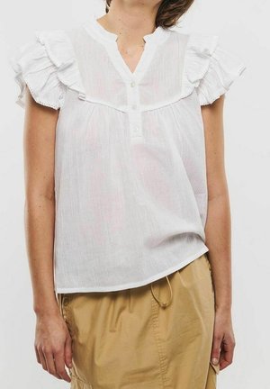 Vrouw in een witte, rimpelige blouse met korte mouwen en een beige rok, staand tegen een effen lichte achtergrond.
