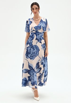 Femme portant une robe longue portefeuille beige avec un grand imprimé floral bleu, des manches courtes volantées, un collier chaîne doré et des talons pointus blancs.