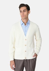 Cardigan bianco lavorato a maglia con scollo a V, bottoni in legno e due tasche frontali; abbinato a una camicia azzurro chiaro e pantaloni grigi.