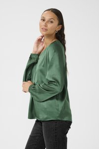 Grüne Satinbluse mit langen, elastischen Ärmeln und V-Ausschnitt. Der Stoff hat eine glatte, glänzende Textur. Getragen mit dunklen Jeans.