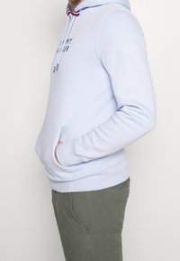 Sudadera azul claro con un bolsillo frontal, que presenta texto negro y un cuello a rayas. La tela parece suave y elástica.