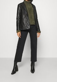 Blazer de cuero negro sobre un jersey de cuello alto de forro polar verde oscuro, combinado con jeans negros de talle alto y botines negros con tacón.