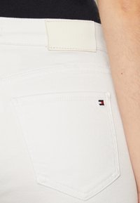 Jeans de mezclilla blanca con un bolsillo trasero que tiene un pequeño emblema en rojo, blanco y azul, y una pequeña etiqueta de cuero beige clara con la marca en la cintura.