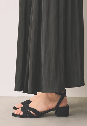 Feet wearing black low-heeled sandals with ankle straps, partially covered by a long, flowing black skirt against a neutral background.