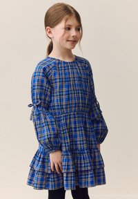 Abito a quadri blu con maniche lunghe a sbuffo, vita arricciata e orlo con volant. Realizzato in tessuto leggero, con dettagli gialli e bianchi.