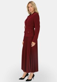 Burgundowa marynarka dopasowana z paskiem i bocznymi kieszeniami, zestawiona z plisowaną spódnicą. Czarne szpilki podkreślają cały strój.