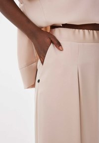 Pantalons plissés beiges avec une taille élastique, des poches latérales et un bouton noir décoratif sur la couture de la poche. Tissu doux et lisse.