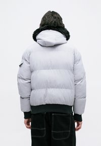 Jachetă puffer în gri deschis, cu glugă pufoasă neagră. Prezintă un tiv și manșete ribbate, cu secțiuni matlasate pentru izolație.