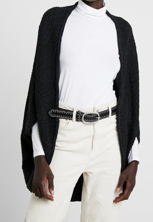 Persona de piel oscura que lleva un suéter de cuello alto blanco, pantalones de pana color crema con cinturón negro con tachuelas y un chal negro texturizado sobre los hombros.