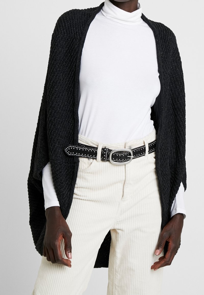 Personne à la peau foncée portant un col roulé blanc, un pantalon en velours côtelé crème avec une ceinture noire cloutée et un châle noir texturé drapé sur les épaules.