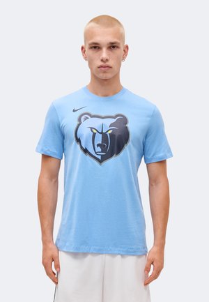 Junger Mann mit kurzen blonden Haaren, der ein hellblaues T-Shirt mit großem Memphis Grizzlies Bärenlogo und weißen Shorts trägt.