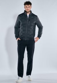 Schwarze gesteppte Jacke mit hohem Kragen, Reißverschlusstaschen und matter Oberfläche, kombiniert mit dunkler Hose und weißen Turnschuhen.
