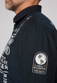 Camisa de manga larga azul marino con tejido texturizado, que presenta texto impreso en blanco y un parche bordado circular del globo en la manga izquierda.