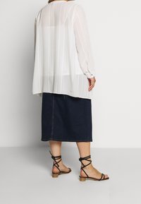 Donna che indossa una blusa bianca trasparente a maniche lunghe, una gonna midi blu scuro e sandali neri a tacco basso con strappi, in piedi contro un muro bianco.