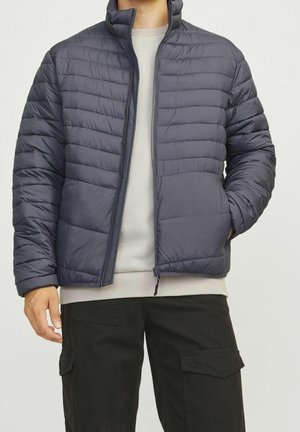 Mężczyzna w ciemnoszarej pikowanej kurtce na jasnoszarym swetrze i czarnych spodniach cargo, stojący z jedną ręką w kieszeni.