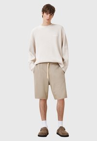 Suéter de punto crema con mangas largas, combinado con shorts beige con cordón ajustable. Zapatos marrones tipo slip-on y calcetines blancos.