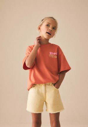 Ung pige med blondt hår iført en orange t-shirt med teksten "good vibes" og gule shorts, stående med den ene hånd i lommen og den anden på nakken.