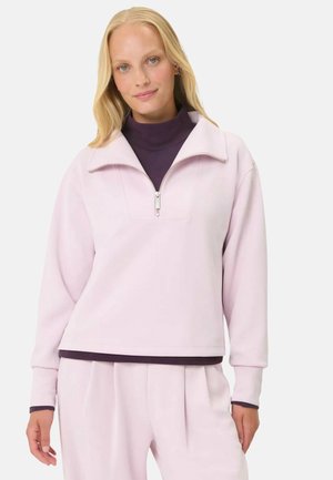 Vrouw draagt een lichtroze sweatshirt met rits over een donkerpaarse coltrui en bijpassende lichtroze broek, staand tegen een witte achtergrond.