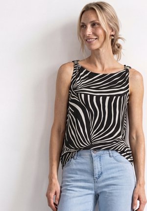 Kvinde med blondt hår smilende, iført sort-hvid zebra-stribet ærmeløs top og lyseblå jeans, stående mod hvid baggrund.