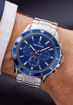 Montre en acier inoxydable argentée au poignet, avec un cadran bleu, trois sous-cadrans, des index argentés et un bracelet en métal.