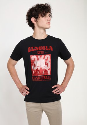 Junger Mann mit lockigem Haar, der ein schwarzes T-Shirt des Manila City 3x3 Basketballturniers trägt, Hände in die Hüfte gestützt, schaut zur Seite.