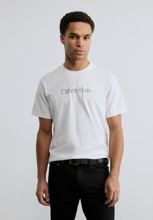 Camiseta blanca de algodón con el logo "Calvin Klein" en gris en el frente, combinada con jeans negros y un cinturón negro, con mangas cortas y cuello redondo.