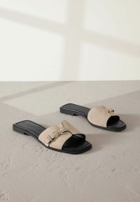 Platta sandaler i beige mocka med en svart lädersula, med ett brett rem och dekorativ detalj i guld.