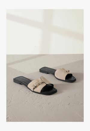 Flache Sandalen aus beigem Wildleder mit einer schwarzen Ledersohle, versehen mit einem breiten Riemen und dekorativem goldfarbigen Hardware-Detail.