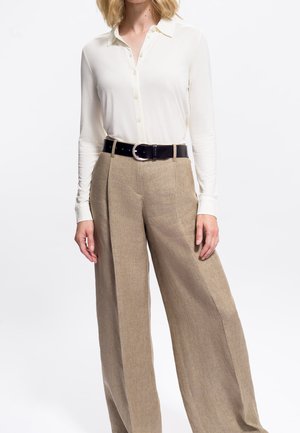 Femme portant une chemise blanche à manches longues boutonnée rentrée dans un pantalon large taille haute beige avec une ceinture noire.