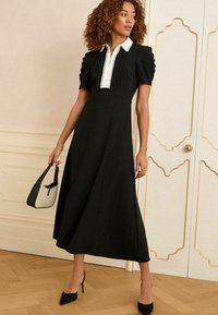 Rochie midi neagră cu mâneci scurte bufante, guler alb și detalii pliate. Femeia ține în mână o geantă mică alb-negru, purtând pantofi cu toc negri.