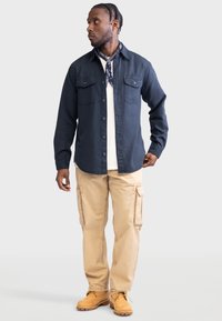Donkerblauwe lange mouwen overhemd met twee borstzakken, gecombineerd met beige cargobroek en tan werkschoenen. Sjaal om de hals geknoopt.