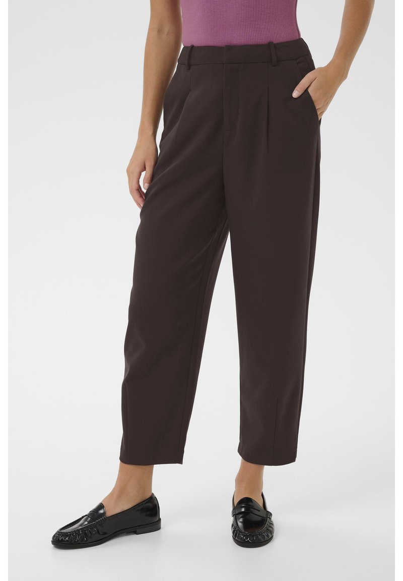 Femme portant un pantalon noir raccourci avec des plis, des mocassins noirs et un haut violet sans manches, debout avec une main dans la poche.