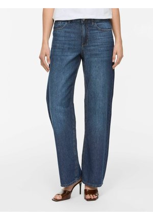 Persona che indossa jeans blu a gamba dritta e sandali marroni con tacco alto e punta aperta, in piedi davanti a uno sfondo chiaro uniforme.