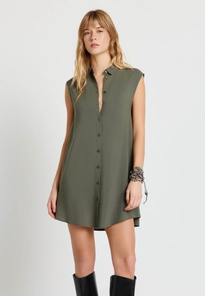 Donna che indossa un abito camicia senza maniche color verde oliva con bottoni, stivali neri a metà polpaccio e braccialetti con perline al polso sinistro, in piedi davanti a uno sfondo bianco.