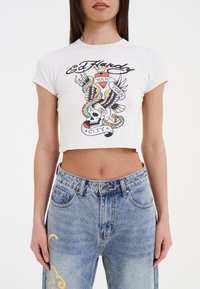 Hvid, kortærmet t-shirt med et farverigt grafisk motiv, der viser designs af et kranium, en ørn og en slange. Sættes sammen med lyseblå denim jeans.