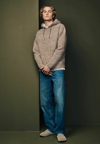 Brun strikket sweater med hætte, parret med blå denim jeans. Personen står mod en grøn baggrund iført beige fodtøj.