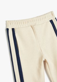 Cremefarbene gerippte Hose mit dunkelblauen Seitestreifen, mit einem gerippten Bund und ohne sichtbare Taschen. Strukturiertes Stoffdetail erkennbar.