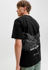 Jaunas vyras, apsirengęs juoda marškinėliais su baltais kontūriniais kalnais ir "JACK & JONES" tekstu ant nugaros, stovi prieš pilką foną.