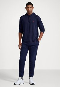 Μπλε σκούρο φούτερ με μακριά μανίκια, τσέπη καγκουρό και μικρό κόκκινο λογότυπο. Συνδυάζεται με αντίστοιχο παντελόνι φόρμας και λευκά αθλητικά παπούτσια.