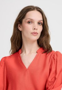 Raudona V formos palaidinė su pūkuotomis rankovėmis, tekstūruota medžiaga ir kuklus apykaklė. Pasižymi laisvu siluetu ir sklandžiu paviršiumi.