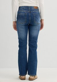 Jeans acampanados de color azul oscuro con un diseño de cinco bolsillos, costuras visibles y una etiqueta de cuero en la cinturilla. La tela parece ser de mezclilla con un ligero desgaste.