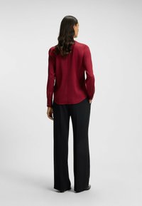 Blusa bordeaux a maniche lunghe con chiusura a bottone sul retro, abbinata a pantaloni neri a gamba larga, realizzati in un tessuto morbido e leggero.
