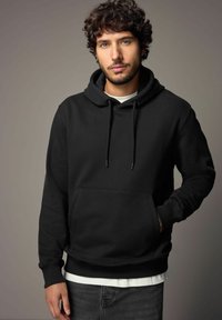 Schwarzer Kapuzenpulli mit Kordelzug und Fronttasche, aus einem weichen Stoff gefertigt. Wird über einem weißen Shirt getragen, das gerippte Bündchen und Saum aufweist.