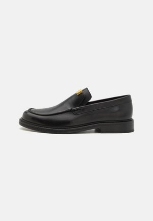 Sapatos de pele preta com um acabamento suave, apresentando um design slip-on e um detalhe em dourado na parte superior. Sola de borracha plana.