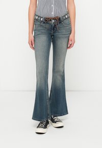 Jeans azuis evasés com um desenho floral bordado no cinto, apresentando um corte de cintura média e lavagem ligeiramente desbotada. Combinados com sapatilhas pretas.