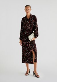 Musta mekko, jossa on punainen ja valkoinen abstrakti kuvio, pitkät hihat ja sivuhalkio, yhdistettynä pieneen vaaleanväriseen clutch-laukkuun ja mustiin korkokenkiin.